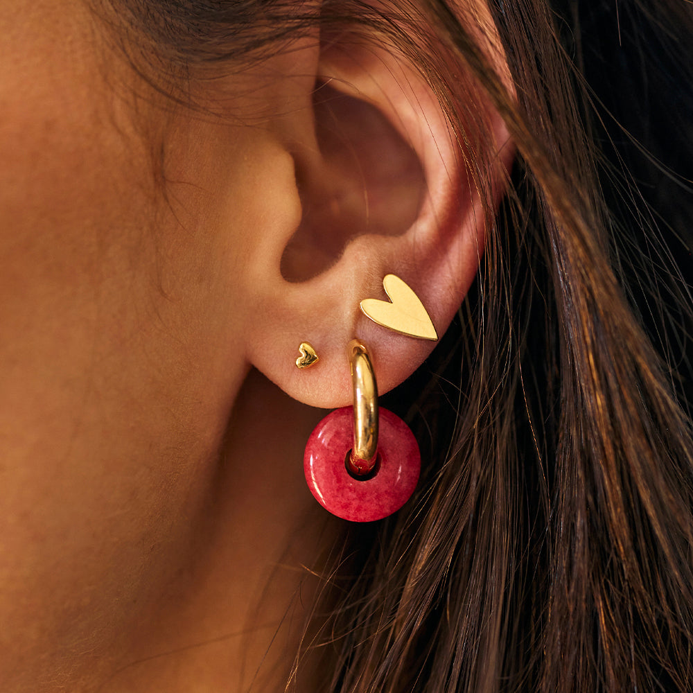 rgola Seven dourada com pingente circular vermelho na orelha, combinada com dois brincos de coração dourados.